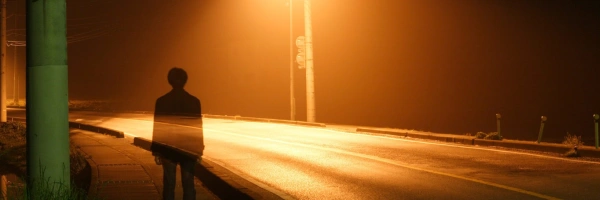 Ein Mann steht im Licht einer Straßenlaterne. Durch ihn schimmert die Straße.