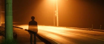 Ein Mann steht im Licht einer Straßenlaterne. Durch ihn schimmert die Straße.