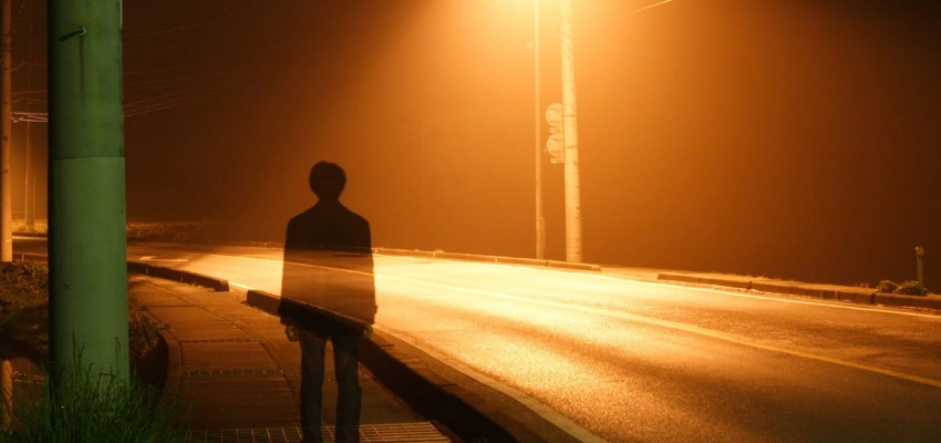 Ein Mann steht im Licht einer Straßenlaterne. Durch ihn schimmert die Straße.