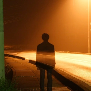 Ein Mann steht im Licht einer Straßenlaterne. Durch ihn schimmert die Straße.