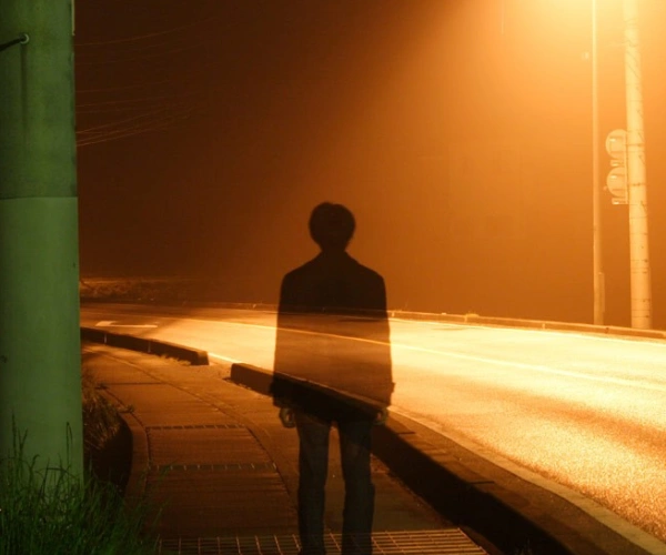 Ein Mann steht im Licht einer Straßenlaterne. Durch ihn schimmert die Straße.