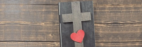 Auf einem, in Leder gebundenem, Buch liegt ein Holzkreuz und ein Herz.