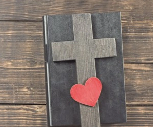 Auf einem, in Leder gebundenem, Buch liegt ein Holzkreuz und ein Herz.