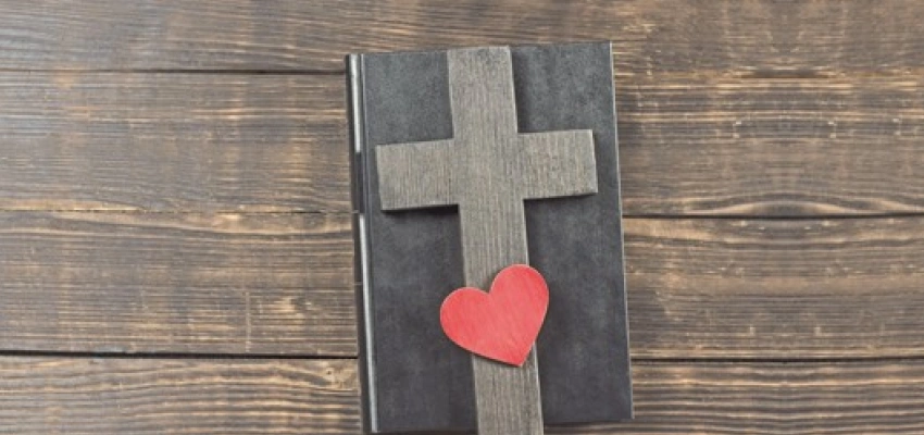 Auf einem, in Leder gebundenem, Buch liegt ein Holzkreuz und ein Herz.