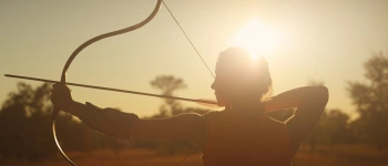 Eine Bogenschützin steht mit gespanntem Bogen in der untergehenden Sonne.