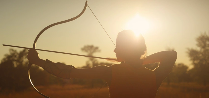 Eine Bogenschützin steht mit gespanntem Bogen in der untergehenden Sonne.