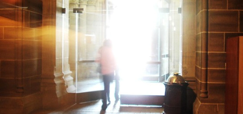 Person, die durch einen Gewölbebogen ins Licht einer offenen Tür tritt.