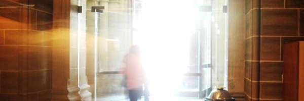 Person, die durch einen Gewölbebogen ins Licht einer offenen Tür tritt.