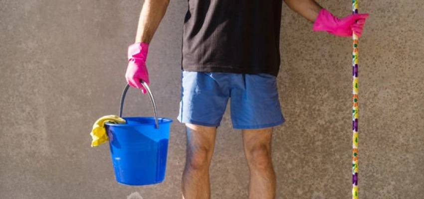 Mensch in kurzer Hose und pinken Gummihandschuhen, mit Putzeimer und Besen in der Hand