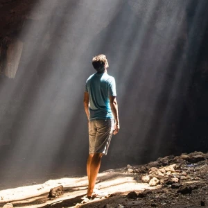 Ein Mann steht in einer Höhle und blickt zu einem Spalt in der Decke, durch den die Sonnenstrahlen fallen.