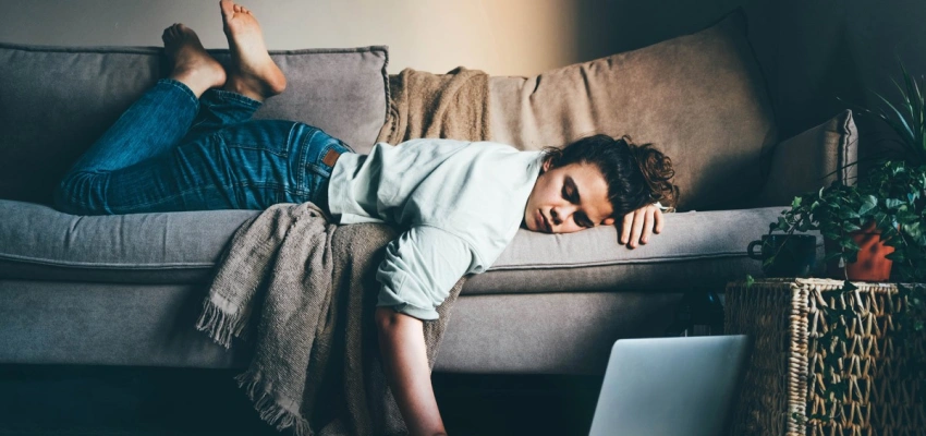 Frau liegt erschöpft auf einem Sofa, eine Hand am Laptop