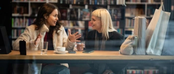 Junge Frauen mit Getränk in engagiertem Gespräch vor Bücherwand